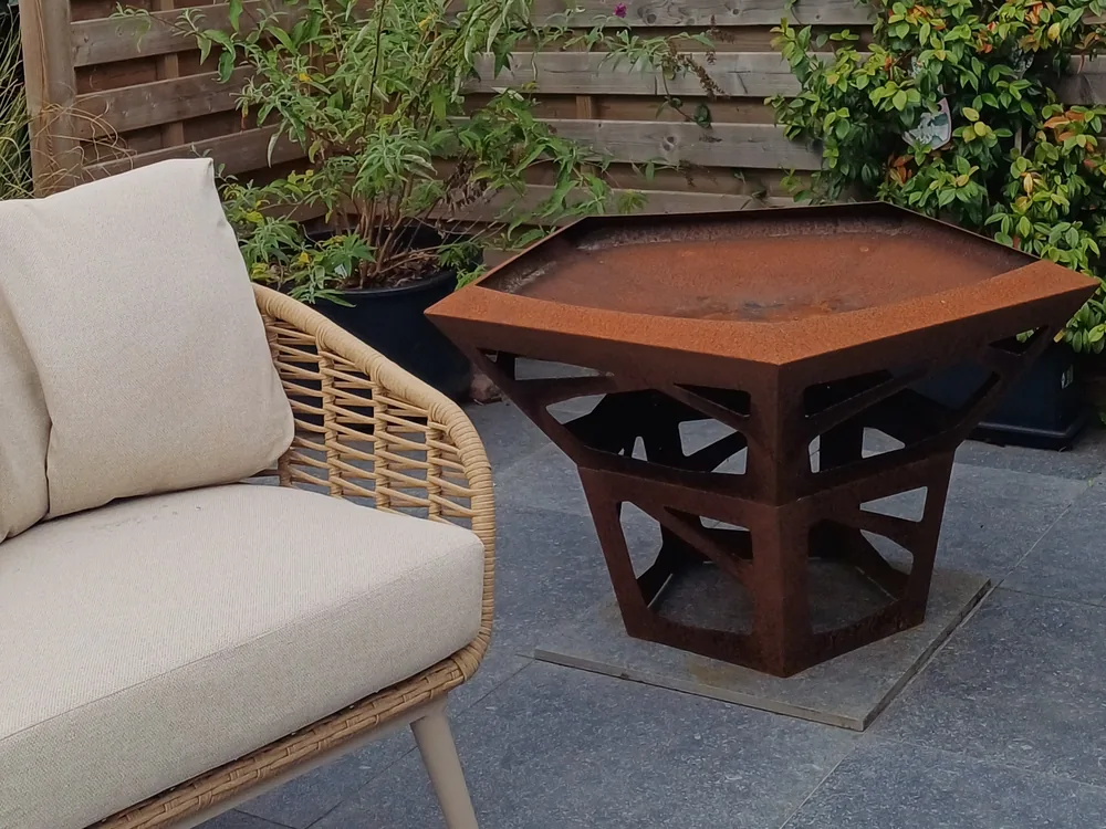 De vuurschaal van cortenstaal op het terras, met een rieten loungebank ernaast en de houten omheining met klimplanten erachter