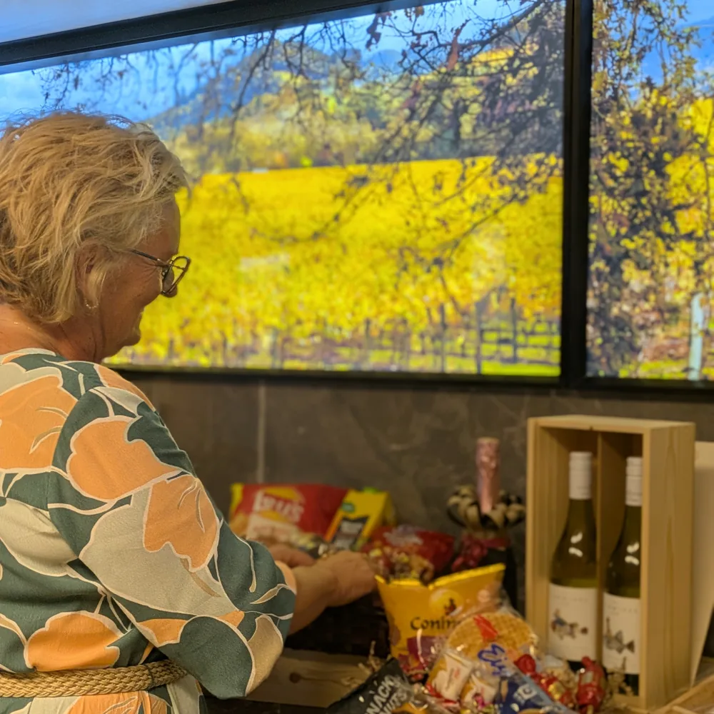 Liliane stelt aan de toonbank een geschenkmand samen, met een houten geschenkdoos met twee flessen wijn ernaast